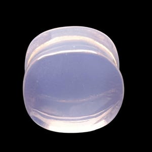 May include: A light blue, translucent ear plug with a rounded shape. The plug has a smooth, polished surface and is set against a black background. The design is simple and minimalist.