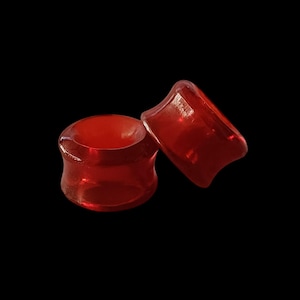 May include: Two red acrylic ear gauges, one slightly larger than the other, against a black backdrop.