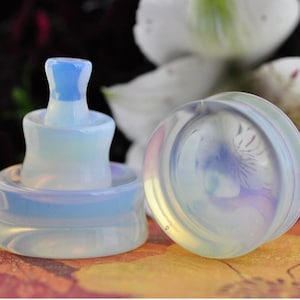 May include: Two iridescent opal ear gauges, one with a layered design and the other with a single round shape. The gauges are a light blue color with a white iridescent sheen.