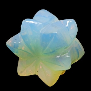 May include: A carved opalescent gemstone in the shape of a starburst flower. The gem has a translucent quality with colors ranging from pale blue to yellow. The object is set against a black background.