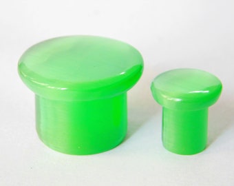 Puede incluir: Dos tapones de botella de vidrio verde, uno más grande que el otro. Ambos tienen una parte superior lisa y redondeada y una base cilíndrica.