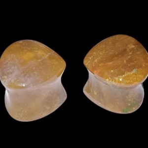 May include: Two teardrop-shaped ear plugs. The plugs are a mix of yellow and brown with a translucent appearance. The base of the plugs is a lighter color than the top. The background is black.