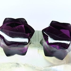 May include: Two polished, star-shaped purple fluorite stones. The stones have a glossy finish and display a range of purple hues, from deep violet to lighter shades. They are set against a white background.