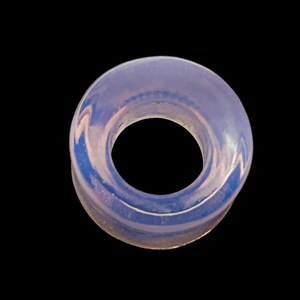May include: A translucent, ring-shaped object with a central hole, set against a black background. The ring has a pale, opalescent quality with hints of blue and white, and appears to be made of glass or a similar material.