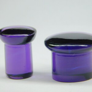 May include: Two purple glass plugs, one is a shorter cylinder shape and the other is a taller cylinder shape.