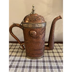 May include: A copper teapot with an ornate floral design and silver accents. The teapot has a spout and a handle and is sitting on a checkered surface.
