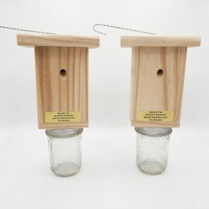 May include: Two wooden bee traps with light-colored pine wood bodies, each featuring a circular entry hole and a clear glass jar attached at the bottom. A metal wire hook extends from the top of each unit. A yellow label reads Best Bee Trap.