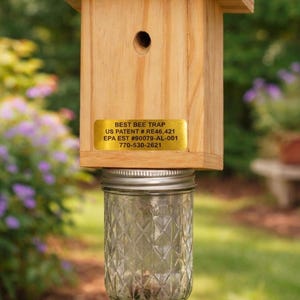Puede incluir: Una trampa para abejas de madera con techo cuadrado y orificio de entrada circular. Una etiqueta dorada dice BEST BEE TRAP, US PATENT # RE46,421, EPA EST #90079-AL-001, 770-530-2621. Un frasco de vidrio transparente con patr&oacute;n de rombos est&aacute; fijado.
