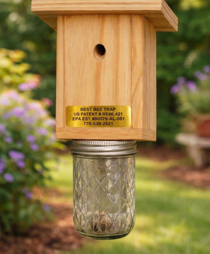 Puede incluir: Una trampa para abejas de madera con techo cuadrado y orificio de entrada circular. Una etiqueta dorada dice BEST BEE TRAP, US PATENT # RE46,421, EPA EST #90079-AL-001, 770-530-2621. Un frasco de vidrio transparente con patr&oacute;n de rombos est&aacute; fijado.