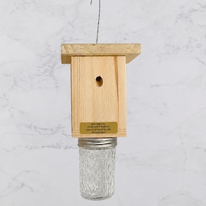 Könnte beinhalten: Eine Bienenfalle aus Holz mit rechteckigem Körper, flacher Oberseite und Metallhaken zum Aufhängen. Ein durchsichtiges, strukturiertes Einmachglas aus Glas ist unten befestigt. Ein goldenes Etikett trägt die Aufschrift: Best Bee Trap, US PATENT # RE46,421, EPA EST # 90079-AL-001, 770-530-2621.