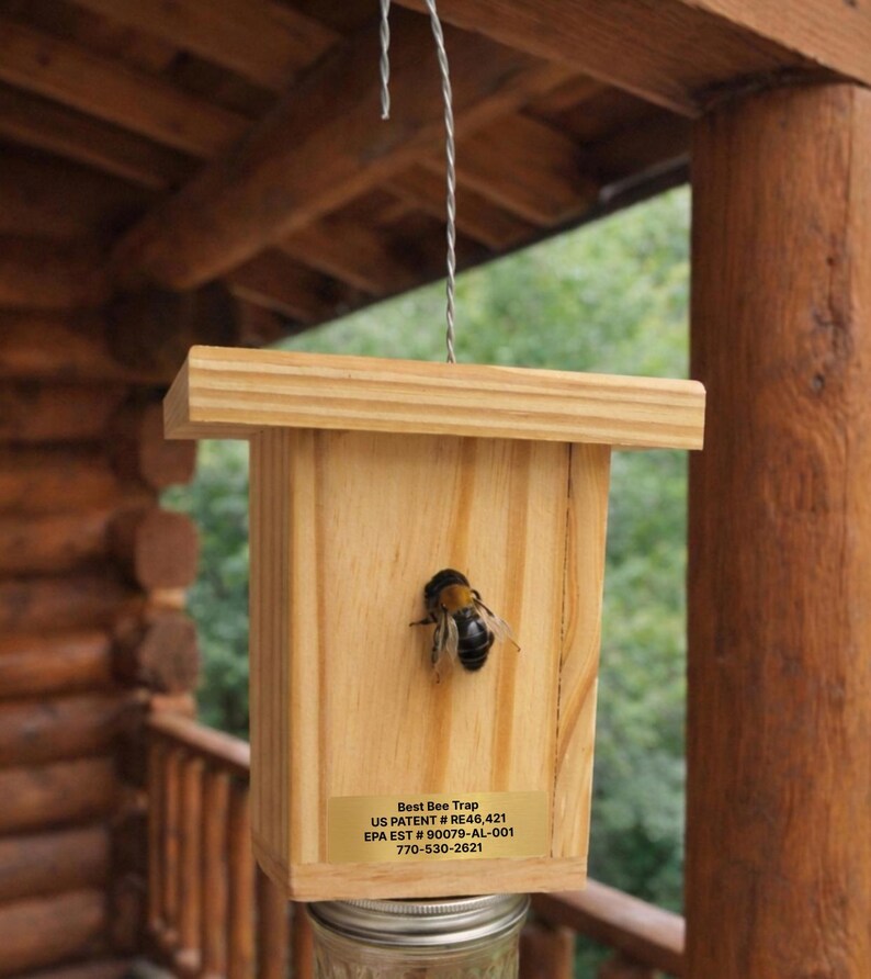 Puede incluir: Una trampa para abejas de madera con un alambre de metal para colgar, con un cuerpo rectangular de madera clara y una etiqueta dorada que dice Best Bee Trap, US PATENT # RE46,421, EPA EST # 90079-AL-001, 770-530-2621, con un abejorro en la parte frontal.