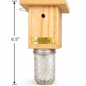 Puede incluir: Una trampa para abejas de madera con un frasco de vidrio transparente unido en la parte inferior. La trampa tiene una etiqueta dorada con el texto BEST BEE TRAP, US PATENT # RE46,421, EPA EST # 90079-AL-001, 770-530-2621. Las dimensiones son 13,3 cm de ancho, 16,5 cm de alto y 29,2 cm de largo con el frasco.