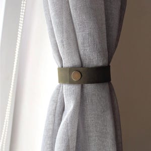 May include: A light gray, semi-sheer curtain held in place by a dark olive green strap. The strap is made of a smooth, matte material and features a single, centered, circular metallic button fastener in a muted brass tone at the front.