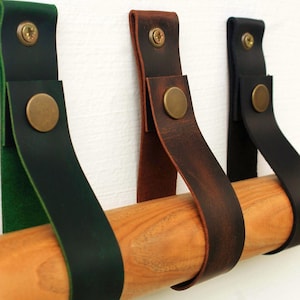 May include: Three leather straps in dark green, brown, and black, each folded and secured with a brass-coloured metal screw and a circular snap button. The straps are looped around a smooth, light-coloured wooden rod, creating a wall-mounted hanger.