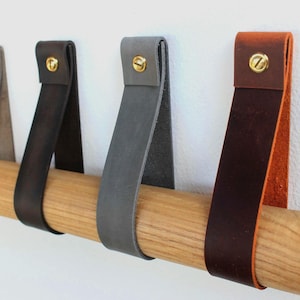 Op de afbeelding: Een reeks lederen ladehandgrepen in verschillende kleuren, waaronder bruin, donkerbruin, zwart en grijs. Elke riem is over een lichtgekleurde houten staaf gevouwen en aan de bovenkant vastgezet met een ronde messingkleurige metalen schroef.