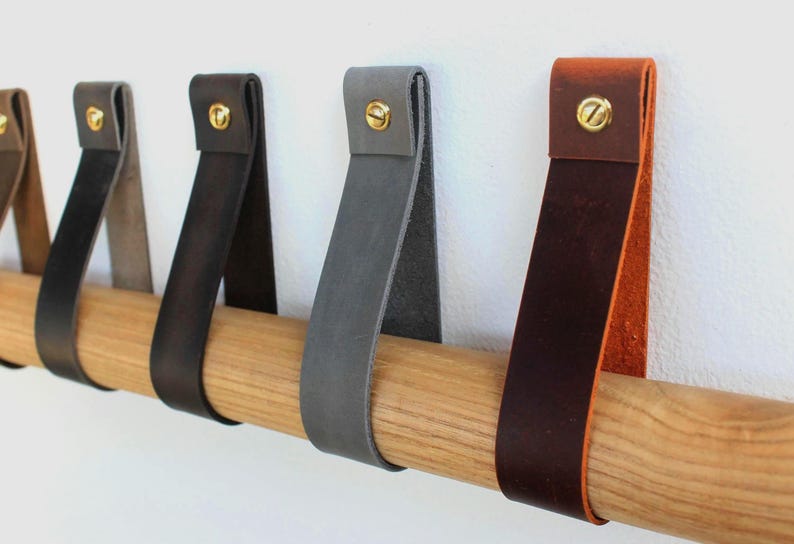 May include: A series of leather drawer pulls in various colours including tan, dark brown, charcoal, and grey, each folded into a loop and secured with a gold-toned metal screw, hanging over a horizontal light-coloured wooden rod.