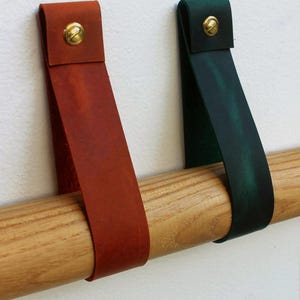 May include: Two leather straps, one in a reddish-brown and one in a dark green, are looped over a light-coloured wooden rod. Each strap is secured at the top with a circular brass-toned metal fastener, creating a simple and functional design.