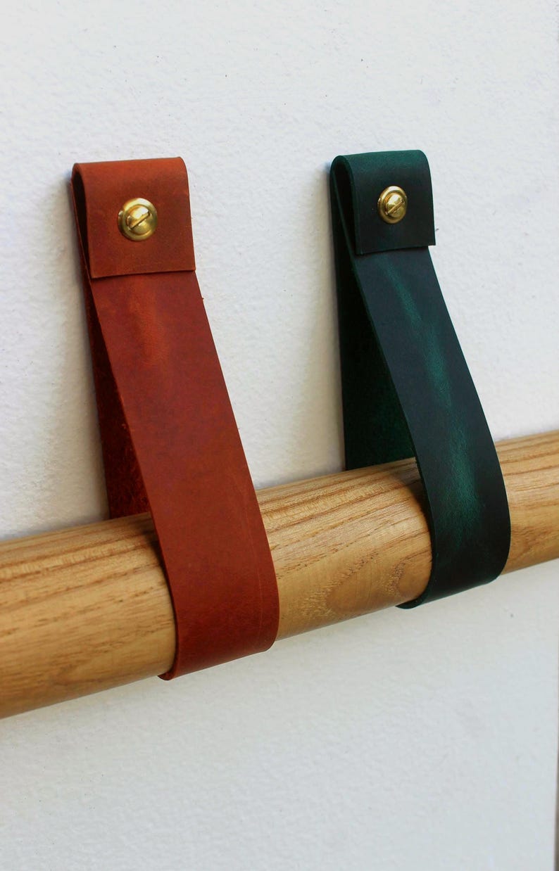 May include: Two leather straps, one in a reddish-brown and one in a dark green, are looped over a light-coloured wooden rod. Each strap is secured at the top with a circular brass-toned metal fastener, creating a simple and functional design.