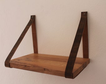May include: A rectangular wooden shelf suspended by two dark brown leather straps. The shelf features a natural wood grain finish and is mounted to the wall with metal hardware at the top of each strap, creating a floating appearance.