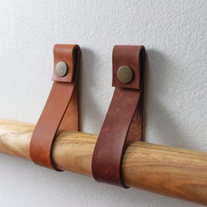 May include: Two leather straps in tan and dark brown colors, each secured with a circular brass-toned metal stud. The straps are looped over a smooth, light-colored wooden rod, creating a functional support system for hanging items.