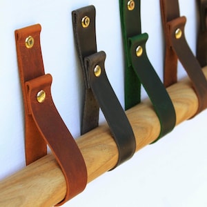 Peut inclure: Une série de lanières en cuir de couleurs fauve, gris foncé, vert et marron, chacune pliée sur une tige en bois cylindrique et fixée avec des vis et des rivets en métal doré. Les lanières sont disposées en rangée, pendant verticalement.