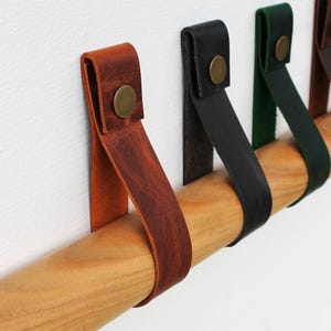Op de afbeelding: Een reeks leren riemen in bruine, zwarte en groene kleuren, elk gevouwen over een cilindrische houten staaf en vastgezet met een ronde, messingkleurige metalen klinknagel. De riemen zijn op een rij gerangschikt en tonen hun verschillende texturen.