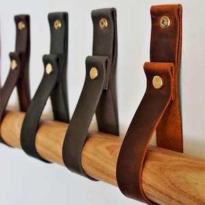 May include: A wooden dowel with several leather straps in varying shades of brown and black. The straps are attached to the wall with gold-coloured rivets. The dowel is a light brown colour.