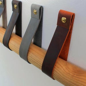 May include: A series of leather straps in various colours including dark brown, grey, and tan, each folded over a cylindrical wooden rod and secured with a gold-toned metal screw. The straps hang vertically from the rod, creating a series of loops.