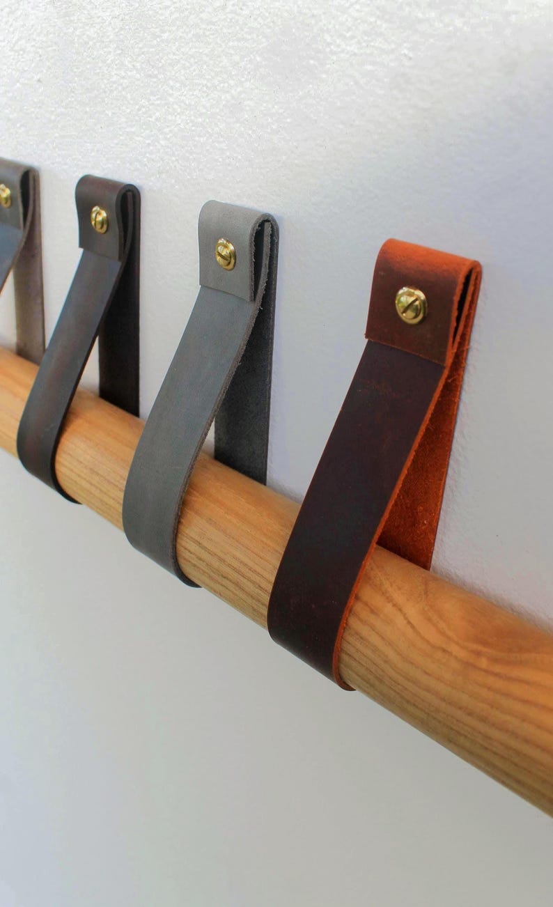 May include: A series of leather straps in various colours including dark brown, grey, and tan, each folded over a cylindrical wooden rod and secured with a gold-toned metal screw. The straps hang vertically from the rod, creating a series of loops.