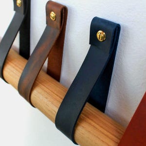 May include: A horizontal wooden rod suspended by multiple leather straps in black, dark brown, and navy blue. Each strap is folded over the rod and secured to the wall with a single brass-coloured metal screw, creating a series of loops.