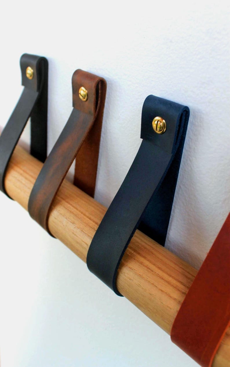 May include: A horizontal wooden rod suspended by multiple leather straps in black, dark brown, and navy blue. Each strap is folded over the rod and secured to the wall with a single brass-coloured metal screw, creating a series of loops.