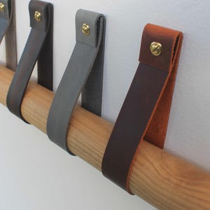 May include: Four leather straps with brass screws are mounted on a wooden dowel. The straps are in shades of brown, grey, and black.