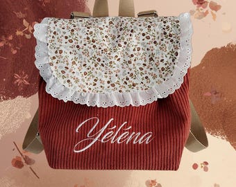 Op de afbeelding: Een roestkleurige rugzak van corduroy met een witte klep met bloemenprint en een geschulpte kanten rand met oogjes. De naam Yélena is in wit cursief schrift op de voorkant geborduurd. De tas heeft beige verstelbare schouderbanden.