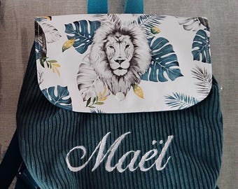 Op de afbeelding: Teal corduroy rugzak met een flap met een leeuwenillustratie en tropische bladeren. De rugzak heeft teal bandjes en een teal handvat. De naam "Maël" is in het wit geborduurd.