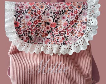 Op de afbeelding: Een oudroze rugzak van corduroy met een bloemenprint op de klep en een witte kanten rand met oogjes. De naam Millie is in wit schrift op het voorvak geborduurd. De tas is voorzien van een handvat en bijpassende roze schouderbanden.