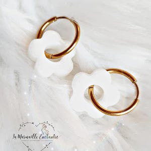 Puede incluir: Un par de pendientes de aro dorados, cada uno con un dije de flor blanca de seis p&eacute;talos. El logotipo de La Marmaille Enchantie, que muestra el contorno de un coraz&oacute;n con una peque&ntilde;a figura en su interior, est&aacute; impreso en negro abajo a la izquierda.