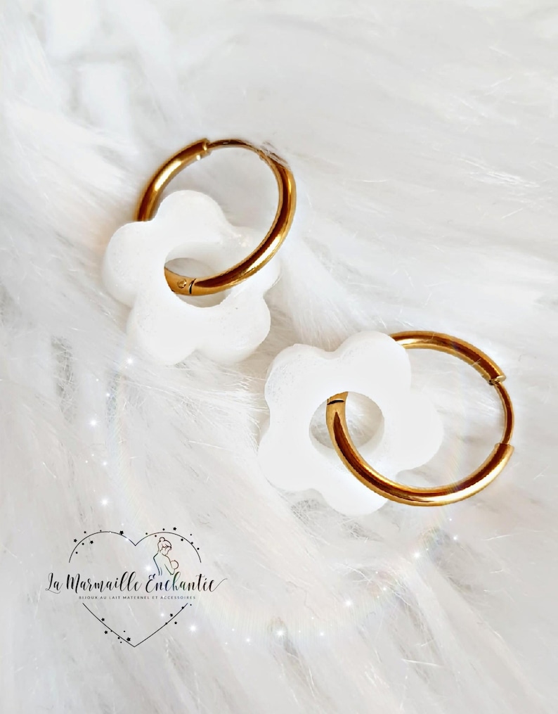Puede incluir: Un par de pendientes de aro dorados, cada uno con un dije de flor blanca de seis p&eacute;talos. El logotipo de La Marmaille Enchantie, que muestra el contorno de un coraz&oacute;n con una peque&ntilde;a figura en su interior, est&aacute; impreso en negro abajo a la izquierda.