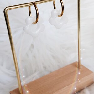 Puede incluir: Un par de pendientes de aro dorados con dijes blancos en forma de flor. Los pendientes se muestran en un soporte de metal dorado con una base de madera. El logotipo La Marmaille Enchant&eacute;e es visible en la esquina inferior derecha.