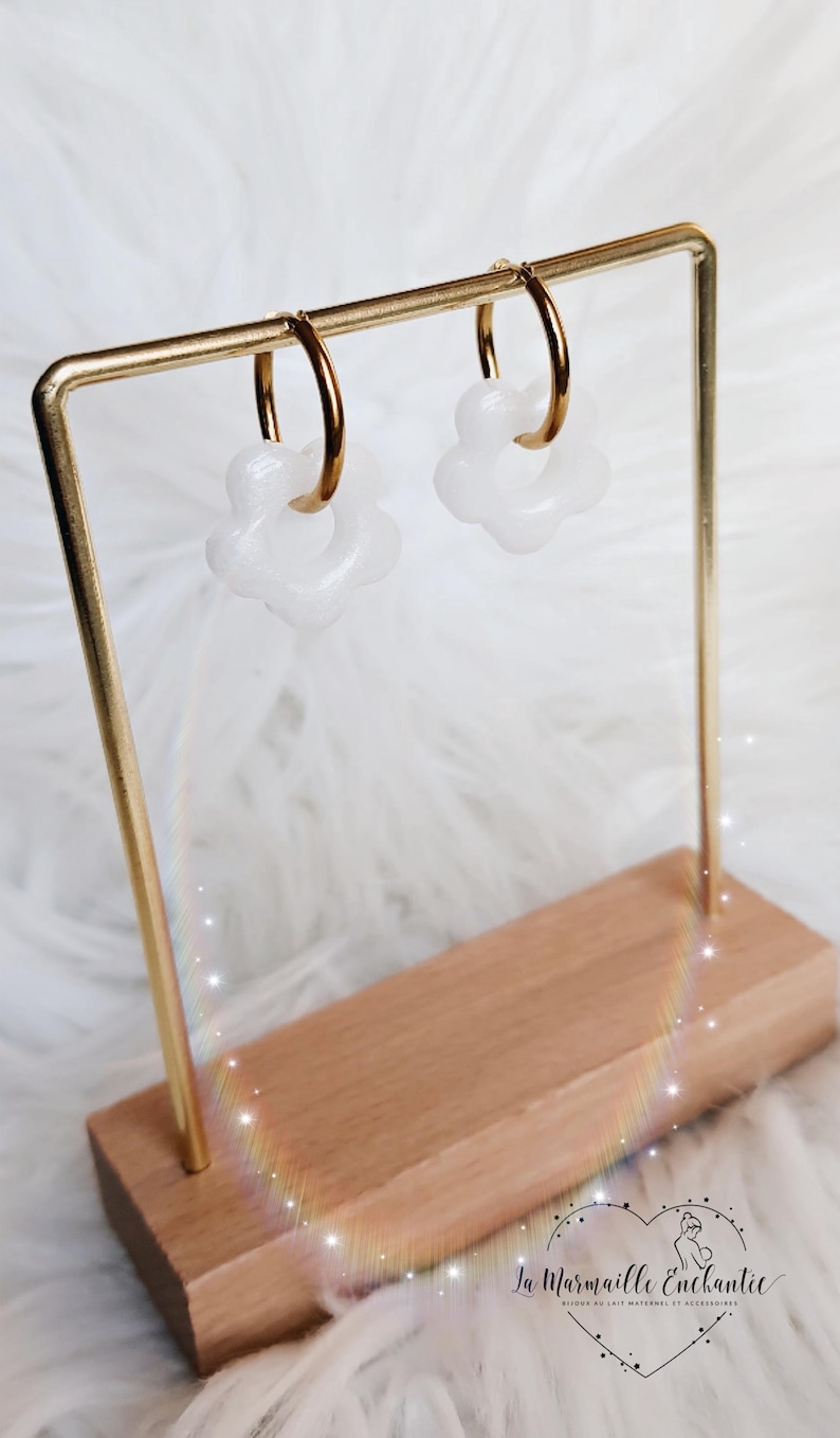 Puede incluir: Un par de pendientes de aro dorados con dijes blancos en forma de flor. Los pendientes se muestran en un soporte de metal dorado con una base de madera. El logotipo La Marmaille Enchant&eacute;e es visible en la esquina inferior derecha.