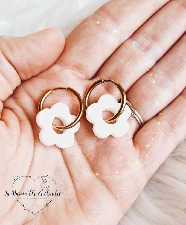 Puede incluir: Un par de pendientes de aro en tono dorado, cada uno con un peque&ntilde;o dije en forma de flor de color blanco mate. Los pendientes tienen un dise&ntilde;o circular con un cierre de bisagra simple, y los dijes florales blancos cuelgan de los aros finos.