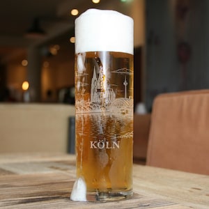 May include: A tall glass of beer with a thick white head. The glass is etched with a detailed cityscape design and the word "KÖLN" at the bottom. The beer is a light amber color, and the glass is sitting on a wooden table.