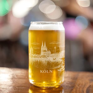 May include: A clear glass beer can-shaped glass filled with light-colored beer and foam. The glass features a white etched design of a cityscape, including a cathedral, bridge, and the word "KÖLN". The glass sits on a wooden surface.