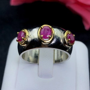 May include: A silver and gold ring with three pink oval-shaped gemstones. The ring is set on a white display stand.