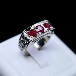 May include: A silver ring with three red gemstones set in a rectangular band. The band has an intricate design.