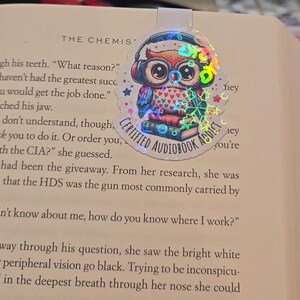 May include: A holographic bookmark featuring a cartoon owl wearing headphones and glasses, perched atop books. The owl has a heart-patterned chest and the text "CERTIFIED AUDIOBOOK ADDICT" is visible.