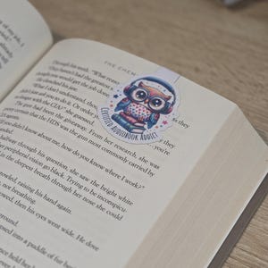 May include: A colorful owl bookmark with headphones, hearts, and the text "Certified Audiobook Addict" is placed on an open book. The bookmark is a sticker with a blue, red, and white color scheme.
