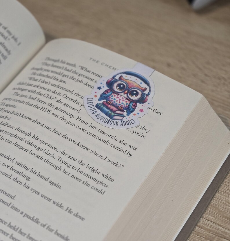 May include: A colorful owl bookmark with headphones, hearts, and the text "Certified Audiobook Addict" is placed on an open book. The bookmark is a sticker with a blue, red, and white color scheme.