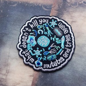 May include: A circular embroidered patch in black and white, featuring the text "What doesn't kill you mutates and tries again." The design includes blue and teal microbe illustrations.