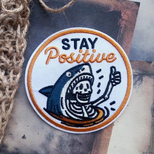 May include: A circular embroidered patch with a white background and an orange border. It features a blue shark with a skeleton torso giving a thumbs up. The text "STAY" is in black and "Positive" is in orange script above the shark graphic.