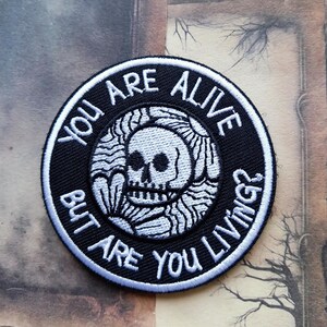 May include: A circular embroidered patch featuring a black background with a white border. The centre displays a white skull graphic surrounded by stylised wave patterns. The text reads: YOU ARE ALIVE BUT ARE YOU LIVING? in white capital letters.